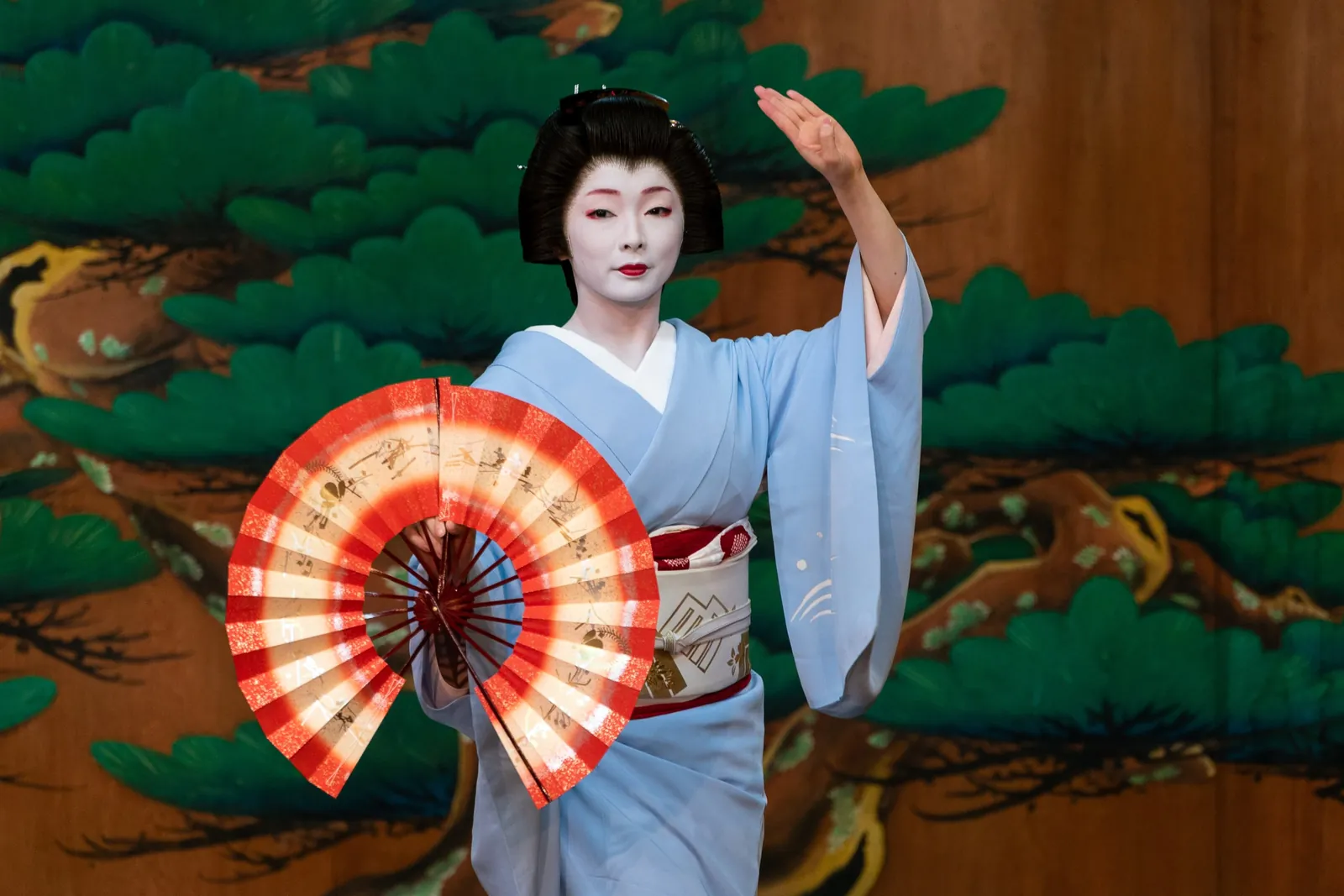 A geiko in a pale blue kimono dances on a stage in front of a painted pine screen, one hand raised and a red and white fan open in the other