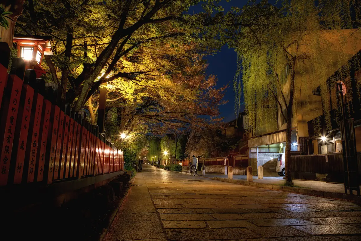A narrow street in Gion at night, lit by lanterns and wet stone, with traditional wooden buildings and willow trees