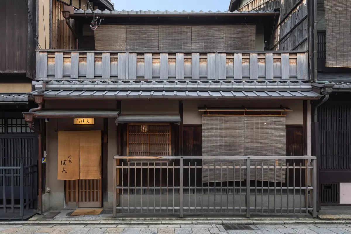 A traditional wooden townhouse facade in Gion with bamboo blinds, wooden balustrades and a yellow lamp