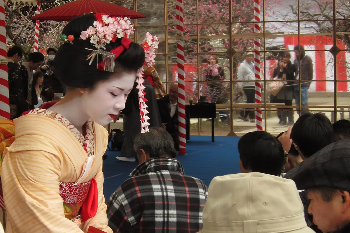 A maiko in a yellow striped kimono with a pink flower hair ornament sets a tea bowl on a white table in front of a seated visitor in a sun hat at Kitano Tenmangū