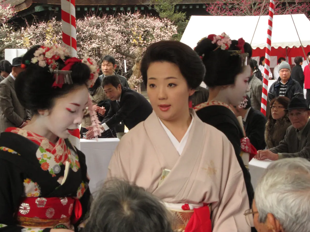 A geiko in a pale pink kimono stands between two maiko wearing elaborate flower hair ornaments at the plum festival at Kitano Tenmangū, with visitors seated behind