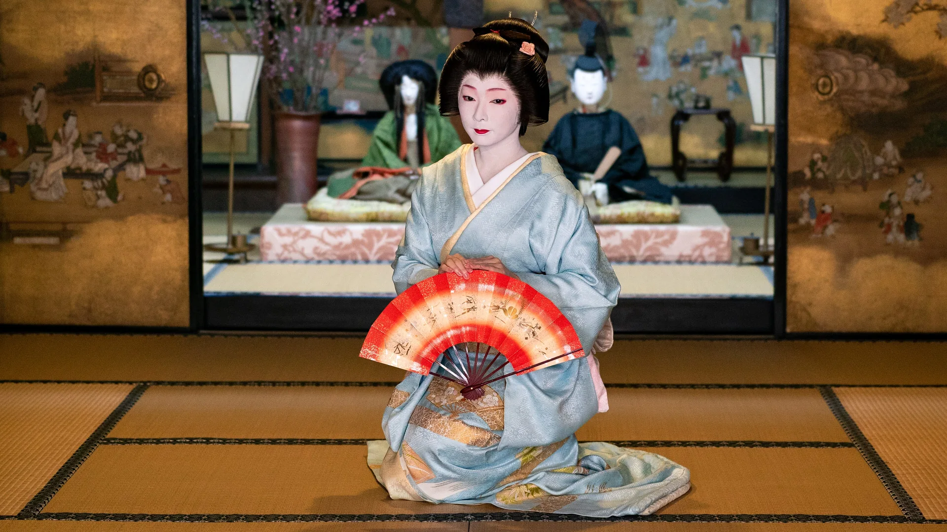 A geiko in a pale blue kimono kneels on a tatami floor in front of a gold-leaf screen, holding a red and white folding fan before her; a general photograph of a geiko's dance, not of this show